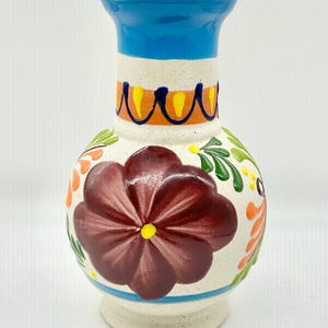 May include: A hand-painted ceramic vase with a bulbous body and a flared neck. The vase features a large maroon flower, orange and green floral accents, and blue and yellow decorative bands. The base is a speckled beige color.