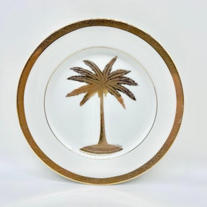 May include: A round white porcelain plate featuring a central metallic gold palm tree illustration on a small island base. The plate has a wide, decorative gold rim with an intricate repeating pattern along the edge and a thin gold inner circle.