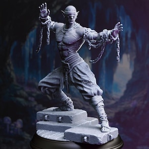 May include: A gray statue of a muscular figure with pointed ears and a blindfold, posed in a dynamic stance. The figure is adorned with chains and stands on a stone platform, set against a dark, cave-like background.