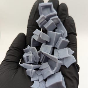 May include: A pile of broken, light gray, plastic pieces in a gloved hand. The pieces are various shapes and sizes, some are square, some are rectangular, and some are irregular.