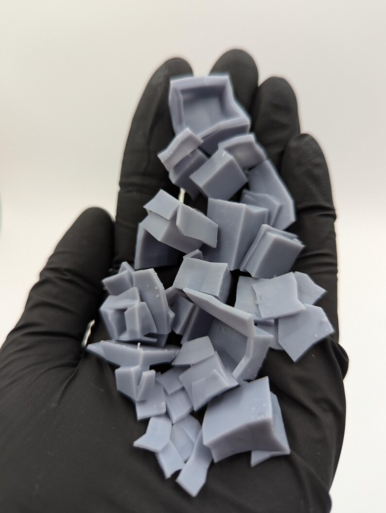 May include: A pile of broken, light gray, plastic pieces in a gloved hand. The pieces are various shapes and sizes, some are square, some are rectangular, and some are irregular.