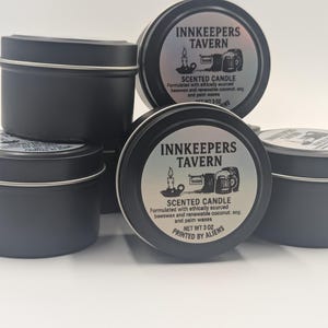 Könnte beinhalten: Vier schwarze Metalldosen mit silbernen Etiketten. Die Etiketten lauten "Innkeepers Tavern" und "Duftkerze". Die Kerzen sind aus ethisch gewonnenem Bienenwachs und erneuerbarem Kokosnuss-, Soja- und Palmwachs hergestellt. Das Nettogewicht beträgt 3 oz. Die Kerzen werden von Außerirdischen gedruckt.