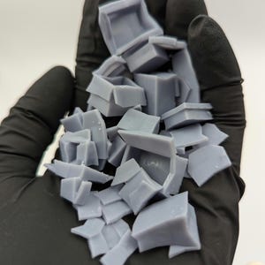 May include: A pile of small, light gray, 3D printed plastic boxes in a gloved hand. The boxes are various sizes and shapes, some are open and some are closed.