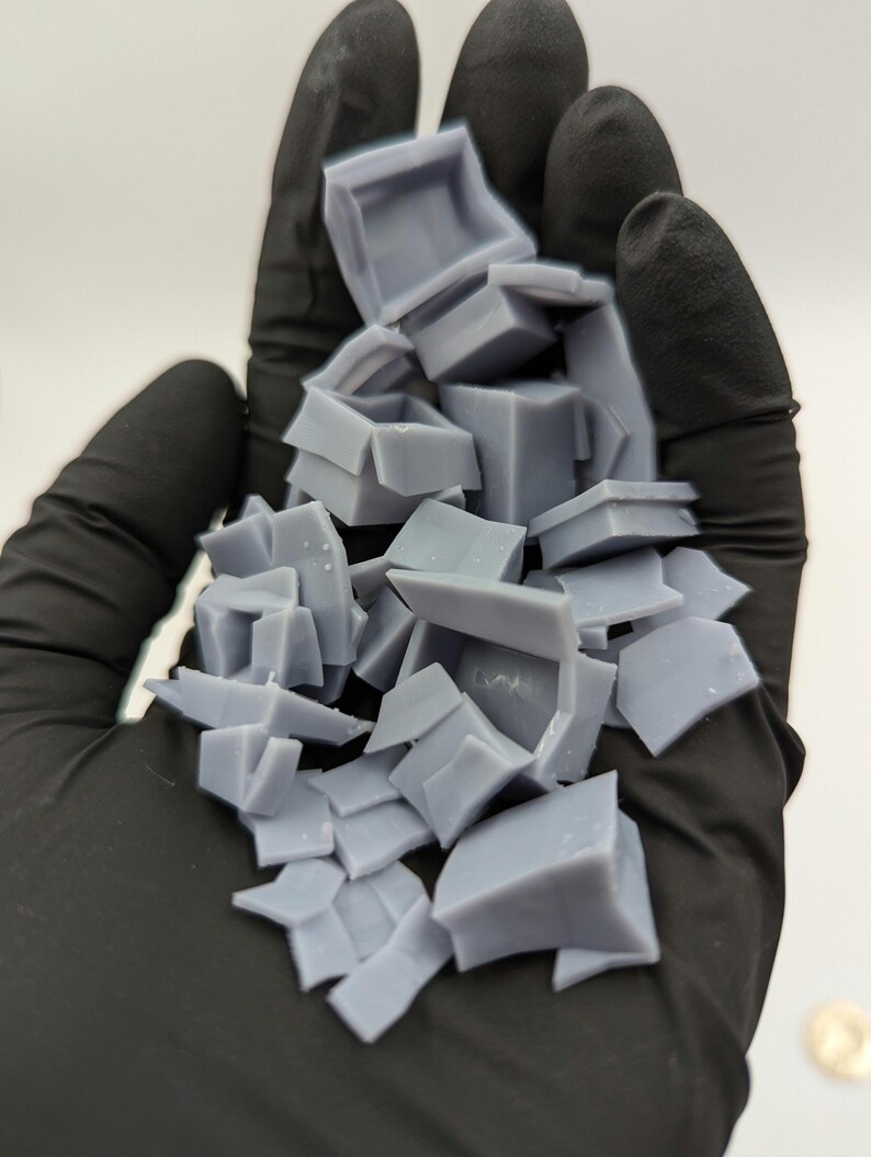 May include: A pile of small, light gray, 3D printed plastic boxes in a gloved hand. The boxes are various sizes and shapes, some are open and some are closed.