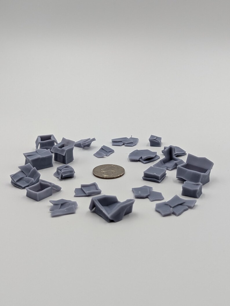 May include: A collection of gray, 3D printed, miniature cardboard boxes in various states of being crumpled and torn. The boxes are arranged in a circle on a white surface.