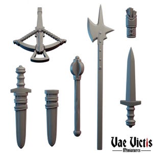 May include: A selection of grey miniature weapons, including a crossbow, axe, mace, dagger, and sword. The weapons are displayed against a white backdrop. The text "Vae Victis - miniatures" is at the bottom right.