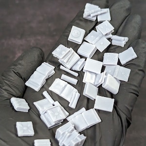 May include: A collection of miniature, white, 3D printed books and scrolls. The books are closed and have various sizes and shapes. The scrolls are rolled up and tied with string.