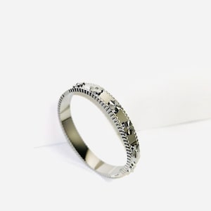 May include: A silver-colored bangle bracelet with a repeating floral pattern. The bracelet has a polished finish and a detailed design. The floral pattern is evenly spaced around the band. The bracelet is displayed on a white surface.