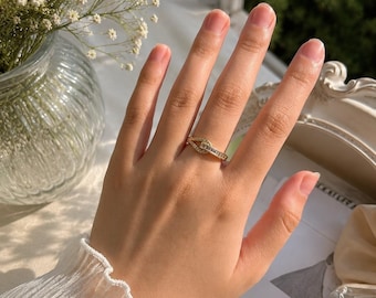 May include: A gold-toned ring with a geometric design, featuring small, clear stones. The ring is displayed on a hand, with a soft, natural background. The image also includes a vase of white flowers and a perfume bottle.