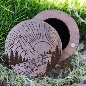 May include: A wooden ring box with a mountain and river scene engraved on the lid. The box is open and the inside is lined with black felt.