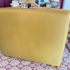 May include: A rectangular mustard yellow hard-shell suitcase with a textured exterior surface. The suitcase features a matching yellow arched top handle and a slim, integrated metal closure mechanism along the upper edge of the main body.
