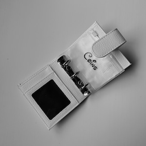 May include: White leatherette binder-style coin purse, open to display clear plastic sleeves and metal rings. The word "Coins" is written in cursive on the plastic. Features a snap closure and a zippered pocket.