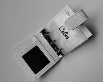 May include: A white faux-leather coin purse, open to show clear plastic sleeves and metal rings. The word "Coins" is written in cursive on the plastic. It has a snap closure and a zipped pocket.