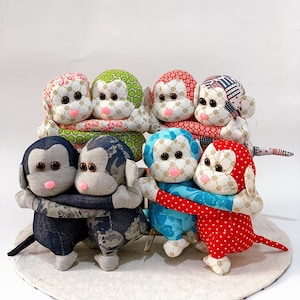 Può includere: Otto giocattoli di scimmia in tessuto fatti a mano, ognuno con un tessuto a fantasia diverso. Le scimmie sono disposte in cerchio, con le braccia l'una intorno all'altra. Le scimmie sorridono tutte e hanno occhi grandi.