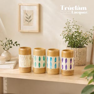 May include: Four wooden vases with a natural finish and decorative bands. Each vase has a unique design: white with a leaf pattern, teal, blue, and purple. The vases are arranged on a wooden shelf with a framed picture and a potted plant.