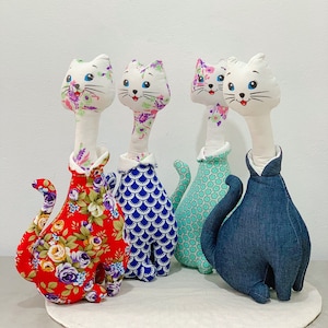 May include: Four handmade plush cat toys with different fabric patterns. The cats are white with black eyes and pink noses. The cats are wearing different outfits, including a floral print, a blue and white scale pattern, a green and white geometric pattern, and a blue denim pattern.
