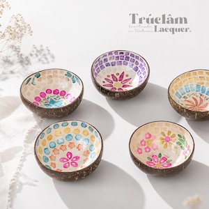 May include: Four small bowls made from coconut shells with dark brown exteriors and interiors decorated with colorful mosaic patterns of flowers and geometric shapes. The text Trúclâm Lacquer is visible in the upper right corner of the image.