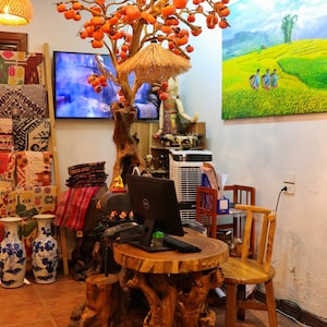 May include: A wooden table with a natural, rough-hewn surface and a unique, branch-like base. The table is decorated with a small potted plant and a laptop computer. A wooden chair with a woven seat sits next to the table. The background features a wall with a painting of a rural landscape and a decorative tree with orange fruit.
