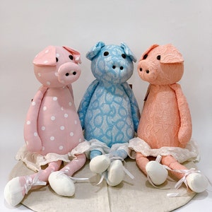 Puede incluir: Tres juguetes de peluche de cerdo hechos a mano. Uno es rosa con lunares blancos, otro es azul con encaje blanco y el último es melocotón con encaje blanco. Los tres tienen un borde de encaje blanco alrededor de la parte inferior y llevan zapatos blancos.