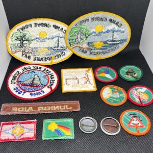 K&ouml;nnte beinhalten: Eine Sammlung von Vintage-Patches und -Anstecknadeln mit verschiedenen Designs und Formen. Die Patches umfassen ovale, rechteckige und kreisf&ouml;rmige Formen mit gestickten Bildern von Segelbooten, B&auml;umen und Text. Die Anstecknadeln sind rund mit Metallr&uuml;cken.