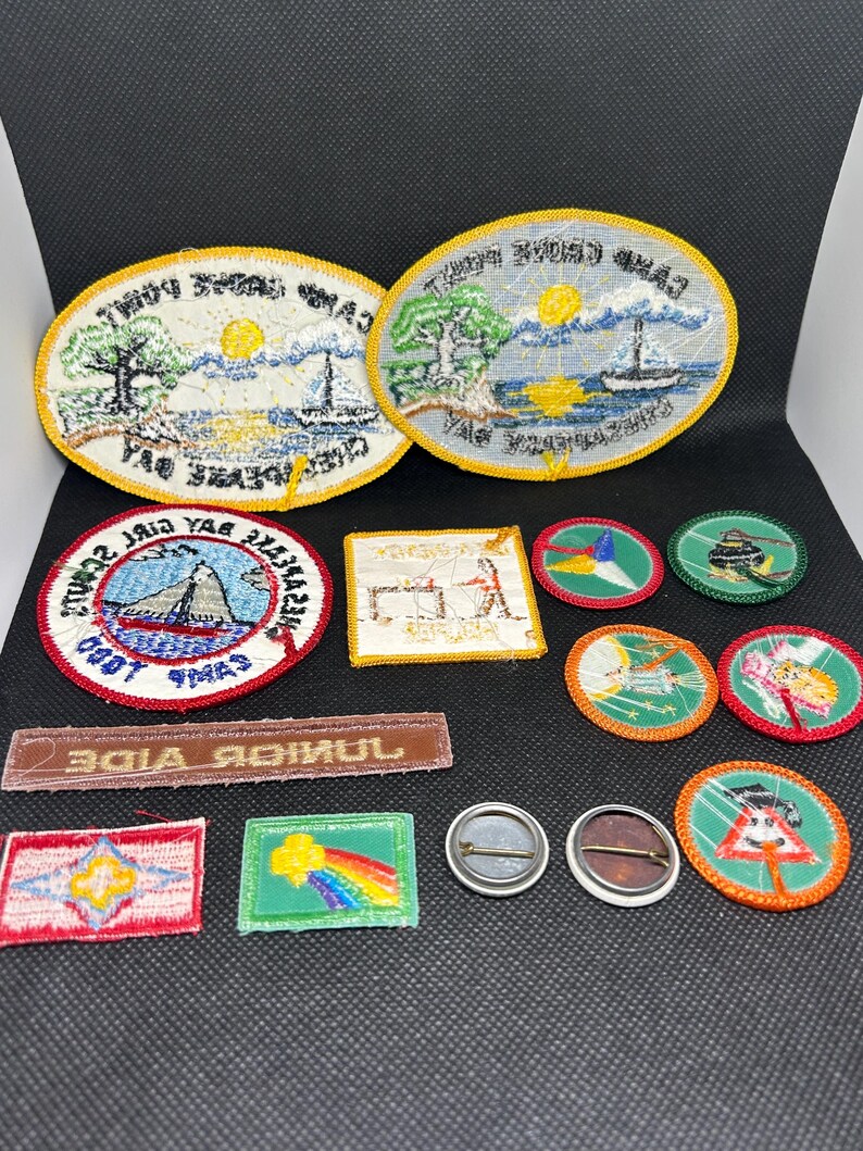 K&ouml;nnte beinhalten: Eine Sammlung von Vintage-Patches und -Anstecknadeln mit verschiedenen Designs und Formen. Die Patches umfassen ovale, rechteckige und kreisf&ouml;rmige Formen mit gestickten Bildern von Segelbooten, B&auml;umen und Text. Die Anstecknadeln sind rund mit Metallr&uuml;cken.