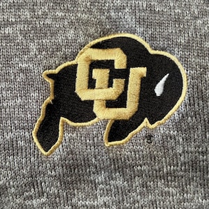 May include: A gray and black embroidered patch with the letters "CU" in gold. The patch is shaped like a buffalo head.