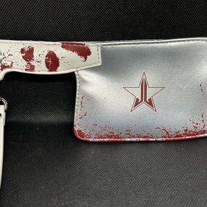 May include: Silver clutch bag with a cleaver design. The handle is white with red splatters, and the body features a red star logo. Includes a white wrist strap and silver hardware.