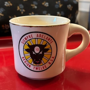 Peut inclure: Une tasse en céramique blanche avec un bord doré et une anse. La tasse présente un emblème circulaire avec une tête de taureau, le texte "SIEMPRE ADELANTE", "REGION TWELVE B.S.A." et le chiffre 12.