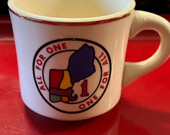 Puede incluir: Taza de cerámica blanca con borde y asa dorados. La taza presenta un gráfico circular con las palabras "ALL FOR ONE" y una ilustración colorida de un contorno estatal con el número "1".