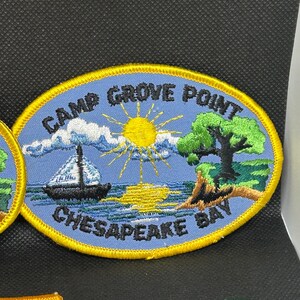 K&ouml;nnte beinhalten: Ovaler, gestickter Aufn&auml;her mit dem Text "CAMP GROVE POINT CHESAPEAKE BAY". Der Aufn&auml;her zeigt ein Segelboot auf dem Wasser, eine Sonne, einen Baum und eine K&uuml;stenlinie. Der Aufn&auml;her hat einen blauen Hintergrund und einen gelben Rand.