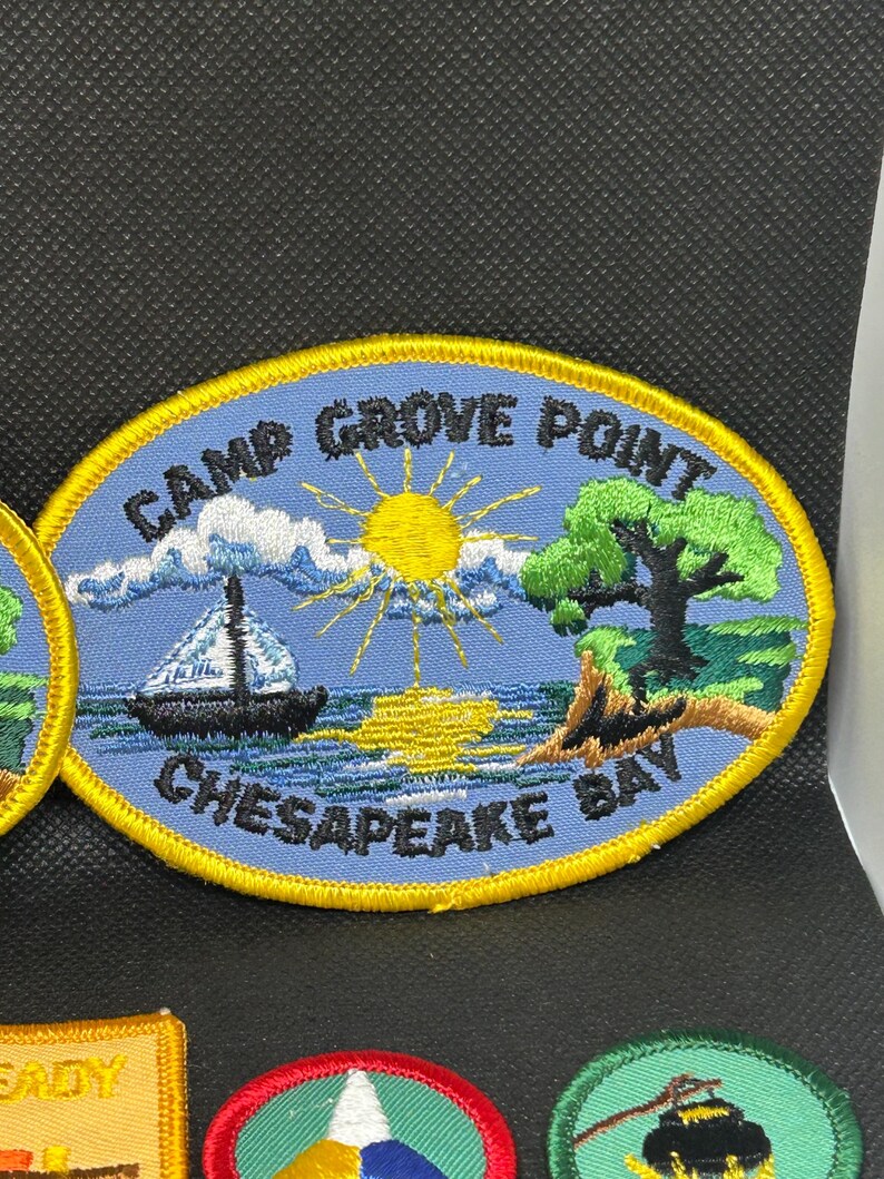 K&ouml;nnte beinhalten: Ovaler, gestickter Aufn&auml;her mit dem Text "CAMP GROVE POINT CHESAPEAKE BAY". Der Aufn&auml;her zeigt ein Segelboot auf dem Wasser, eine Sonne, einen Baum und eine K&uuml;stenlinie. Der Aufn&auml;her hat einen blauen Hintergrund und einen gelben Rand.