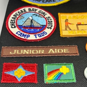 K&ouml;nnte beinhalten: Eine Sammlung gestickter Aufn&auml;her, darunter ein runder Aufn&auml;her mit einem Segelboot und dem Text "Chesapeake Bay Girl Scouts Camp Todd". Weitere Aufn&auml;her zeigen Texte wie "Junior Aide" und ein Regenbogen-Design.