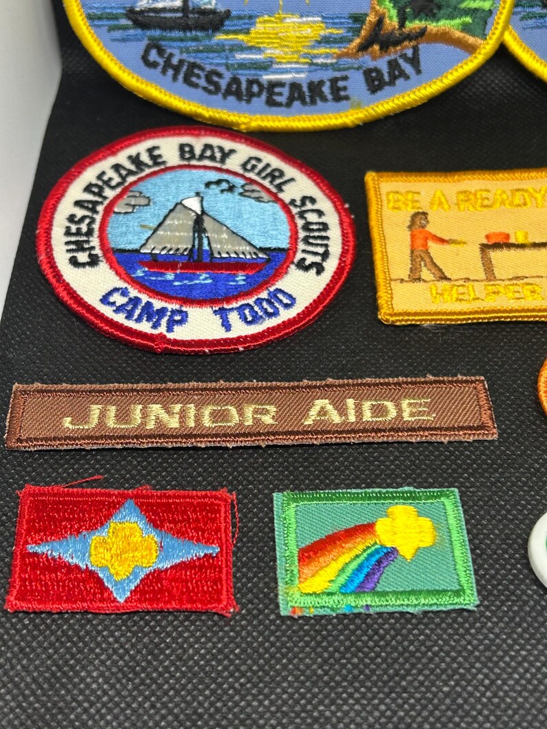 K&ouml;nnte beinhalten: Eine Sammlung gestickter Aufn&auml;her, darunter ein runder Aufn&auml;her mit einem Segelboot und dem Text "Chesapeake Bay Girl Scouts Camp Todd". Weitere Aufn&auml;her zeigen Texte wie "Junior Aide" und ein Regenbogen-Design.