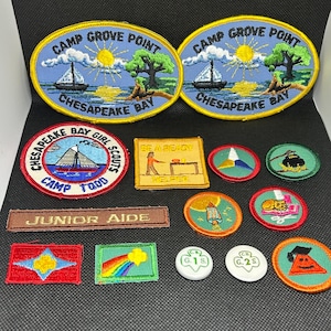 K&ouml;nnte beinhalten: Eine Sammlung gestickter Aufn&auml;her mit verschiedenen Designs. Zwei ovale Aufn&auml;her zeigen "Camp Grove Point Chesapeake Bay" mit einem Segelboot und einem Baum. Weitere Aufn&auml;her enthalten Texte wie "Junior Aide" und "Be Ready Helper" sowie Pfadfinderinnen-Abzeichen.