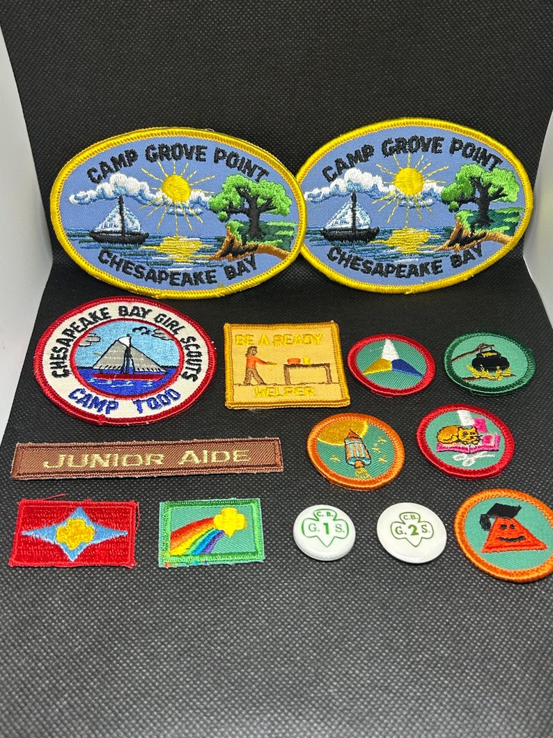 K&ouml;nnte beinhalten: Eine Sammlung gestickter Aufn&auml;her mit verschiedenen Designs. Zwei ovale Aufn&auml;her zeigen "Camp Grove Point Chesapeake Bay" mit einem Segelboot und einem Baum. Weitere Aufn&auml;her enthalten Texte wie "Junior Aide" und "Be Ready Helper" sowie Pfadfinderinnen-Abzeichen.