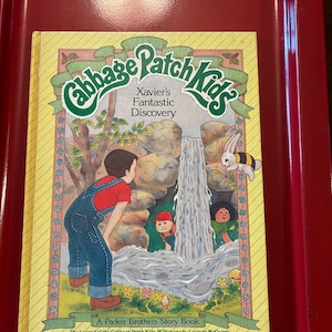 Könnte beinhalten: Ein Vintage-Bilderbuch "Cabbage Patch Kids", "Xavier's Fantastic Discovery", zeigt eine farbenfrohe Illustration von Kindern in der Nähe eines Wasserfalls. Das Cover ist gelb mit grünen und rosa Akzenten, und es ist ein Parker Brothers Story Book.