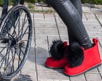 May include: A pair of bright red felt boots with large black fur pom-poms. The boots have a rounded toe and a small heel. The person is wearing black leggings. A vintage black metal wheel is in the background.