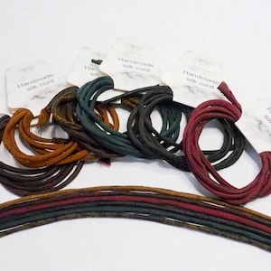 May include: A collection of handmade silk cords in various colors, including brown, orange, green, and burgundy. Each cord is coiled and labeled with a small tag. Several cords are laid out in a row.