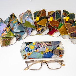 May include: A collection of colorful, patchwork fabric eyeglass cases and matching folded cases. The cases feature a variety of patterns and colors, including floral and geometric designs. A pair of eyeglasses rests below the cases.