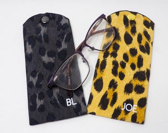 Pode incluir: Dois estojos para óculos de estampa de leopardo com manchas pretas e texto branco. O estojo à esquerda é cinza com o texto "BL" e o estojo à direita é amarelo com o texto "JOE".
