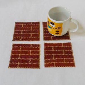 May include: Four brown square coasters with a brick pattern and gold stitching. A white mug with a yellow design sits on top of one of the coasters.