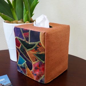 May include: A brown tissue box cover with a patchwork design of colorful fabric squares. The fabric squares are sewn together with gold thread. The tissue box cover is sitting on a wooden table.