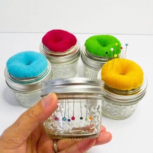 Op de afbeelding: Een verzameling glazen potten met kleurrijke, donutvormige speldenkussens. De speldenkussens zijn in de kleuren rood, groen, geel en blauw. Sommige potten hebben spelden erin. De potten worden in een hand gehouden.