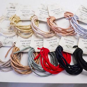 Op de afbeelding: Een verzameling handgemaakte zijden koorden in diverse kleuren: wit, goud, roze, lichtblauw, grijs, bruin, rood, marineblauw en zwart. Elk koord is opgerold en voorzien van een label met "Handmade silk cord".