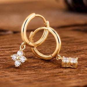 May include: A pair of gold hoop earrings with dangling charms. One earring features a four-petal flower design with clear gemstones, and the other has a rectangular charm with three gemstones. The earrings are displayed on a wooden surface.