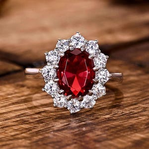 May include: A silver ring featuring a large, oval-cut, deep red gemstone surrounded by a halo of smaller, clear gemstones. The ring is set against a wooden surface, highlighting the jewelry's sparkle and design.