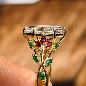 Könnte beinhalten: Ein goldener Ring mit einem großen, rechteckig geschliffenen Edelstein. Das Ringdesign beinhaltet ein Blumenmotiv mit roten und grünen Akzenten. Das Band hat ein verdrehtes, weinrebenartiges Aussehen.
