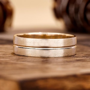 Könnte beinhalten: Ein goldener Bandring mit gebürstetem, mattem Finish und einer einzelnen horizontalen Rille, die um die Mitte der Außenfläche verläuft. Der Ring hat ein flaches Profil und eine gleichmäßige Breite, was einen metallischen Glanz zeigt.