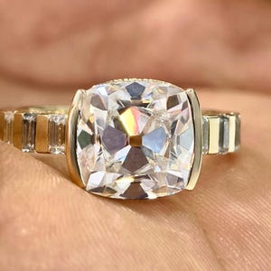 May include: A gold ring featuring a large, square-cut, clear gemstone. The ring's band has a series of rectangular accents on each side of the main stone. The ring is held in a hand, showcasing its brilliance and design.