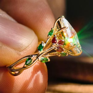 May include: A gold ring featuring a large, pear-shaped gemstone with a rainbow effect. The ring is adorned with smaller green and red gemstones, and has a delicate, leaf-like design on the band.